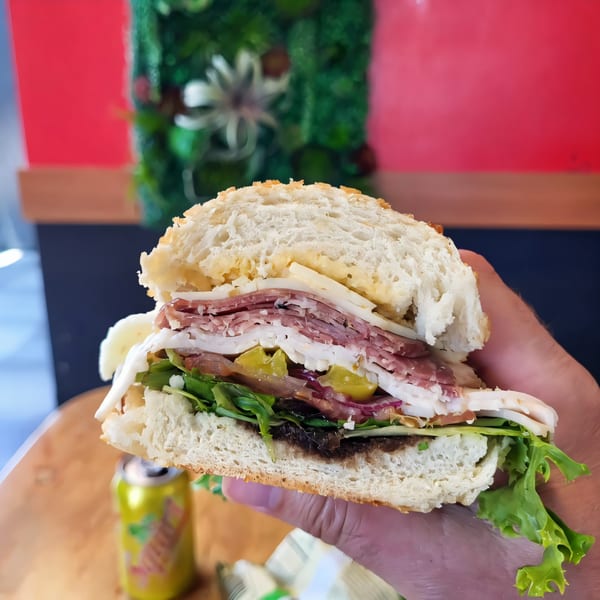 A hand holds a large, fresh sandwich from The Sandwich Spot, filled with meats, cheese, and vegetables.