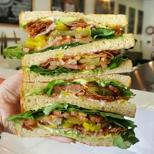 A hand in a clear glove holds a towering, multi-layered sandwich from The Sandwich Spot.