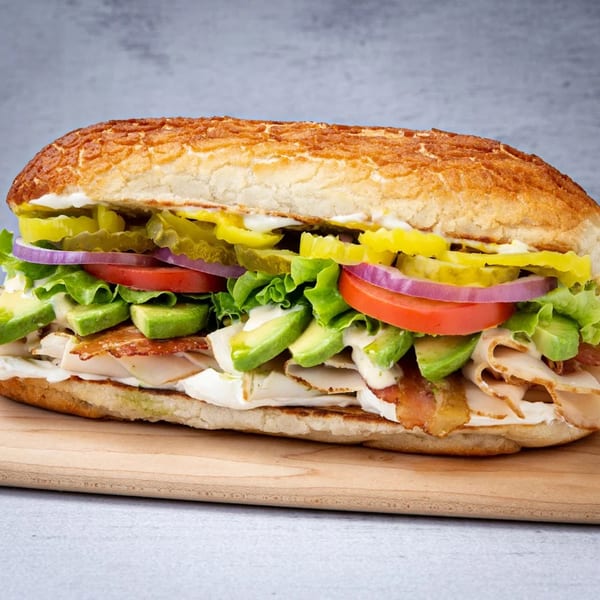 A large, loaded turkey and bacon sandwich with avocado, lettuce, tomato, onion, and pickles.